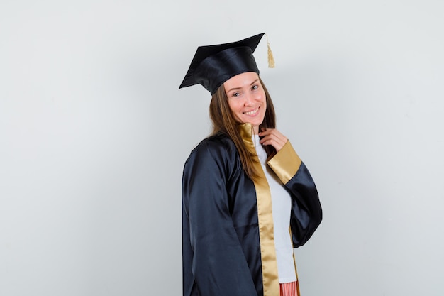 Female graduate posing while standing in academic dress and looking cute , front view.