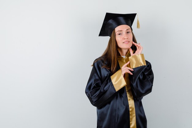 Female graduate posing while standing in academic dress and looking delicate , front view.