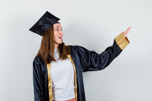 Female graduate pretending to show something in academic dress and looking astonished , front view.