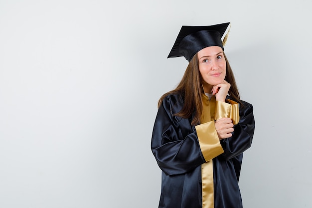 Выпускница, подпирая подбородок рукой в академической одежде и мечтательно выглядя, вид спереди.