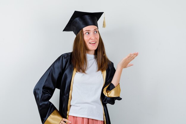 Выпускница-женщина в униформе, повседневной одежде показывает что-то, глядя вверх и весело, вид спереди.