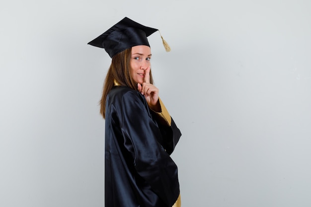 制服を着た女性の卒業生は、沈黙のジェスチャーを示し、賢明に見えます。