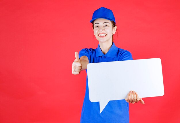 Female guide in blue uniform holding a white rectangular info board and showing enjoyment sign. 