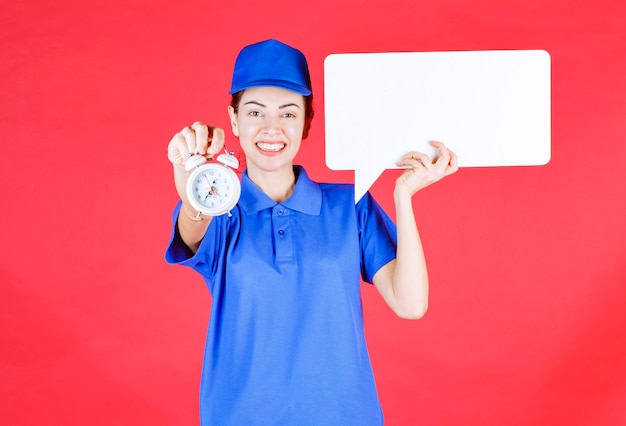 Female guide in blue uniform holding a white rectangular info board with an alarm clock . 