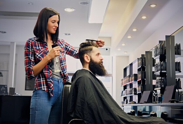 Female hairdresser cutting hair of bearded hipster male in a saloon.