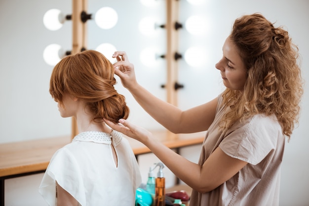 Female hairdresser making hairstyle to redhead woman in beauty salon
