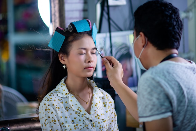 female hairdresser standing, face makeup and making hairstyle to cute lovely young woman in beauty salon