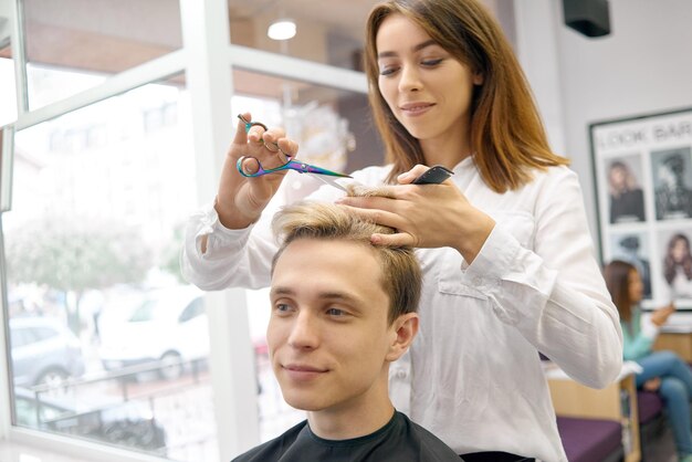 引き締まった髪のハンサムなモデルのためにヘアカットをしている女性のヘアスタイリスト