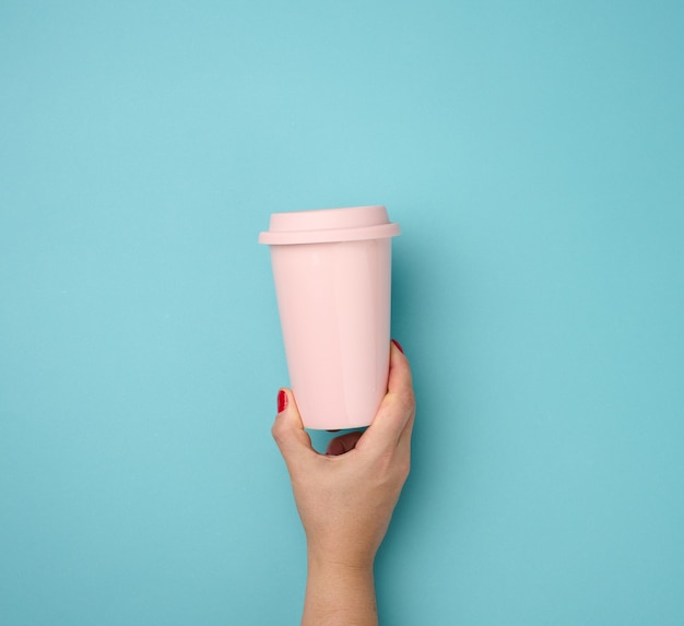 青い​背景​に​プラスチック​の​蓋​が​付いた​ピンク​の​セラミックマグカップ​を​持っている​女性​の​手​、​休憩​時間​と​コーヒー​を​飲む