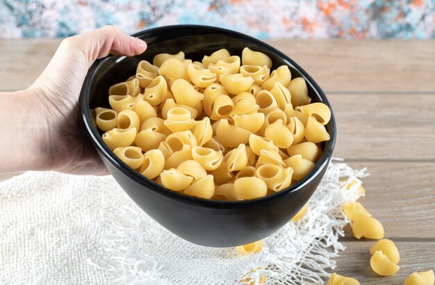 Female hand holding bowl full of dry pasta on wooden table.