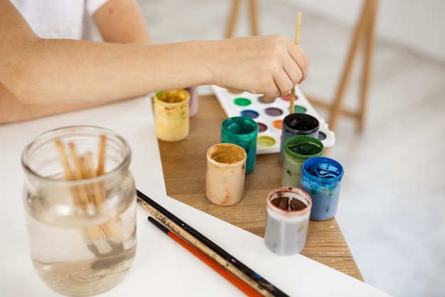 Female hand holding brush and deeping it into paint. Kid painting using watercolour during lesson in the art room.