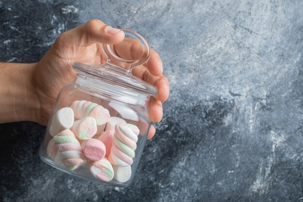 Female hand holding glass jar of colorful marshmallows on marble background