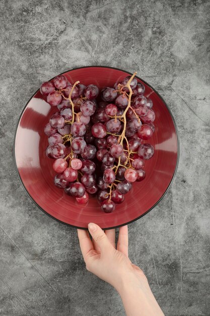 Female hand holding plate of fresh grapes placed on marble surface.