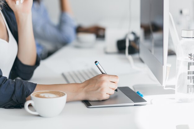 Female hand holding stylus on tablet. Indoor portrait of freelance web-developer working on project during coffee-break in office.