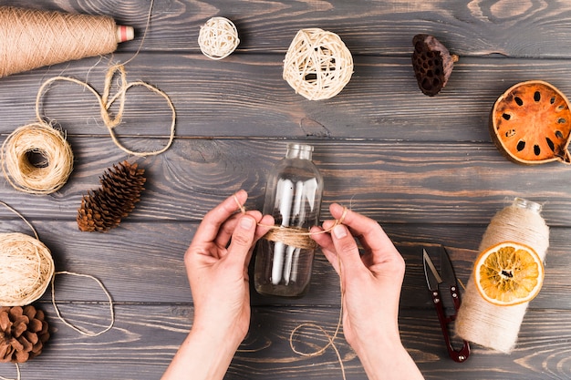 Female hand tying glass bottle with string near cutter; dry lotus pod; dried fruit slices; pine cone over textured wooden surface