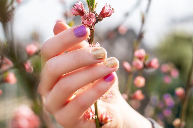 Female hand with gold and purple nail design holding blossom branch.