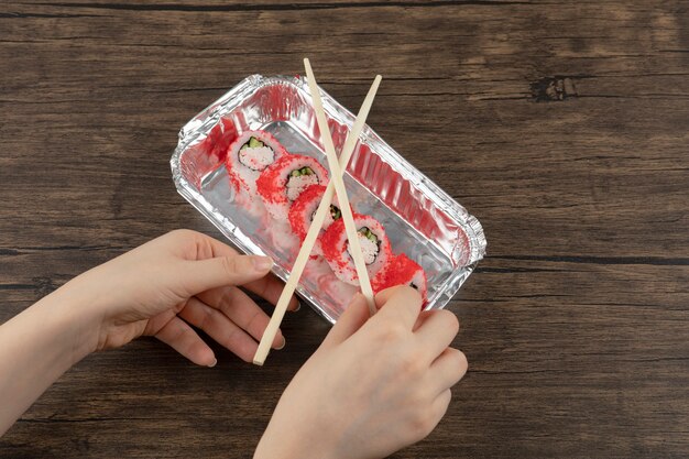 Female hands holding aluminum foil plate of sushi rolls on wooden surface