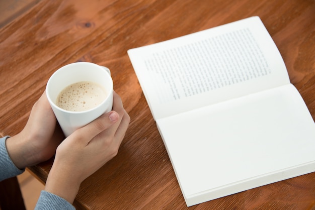 Mani femminili in possesso di tazza di caffè sul tavolo con il libro