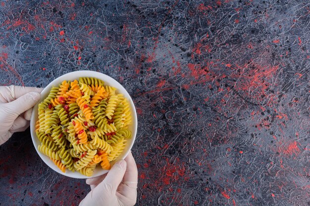 Female hands holding a white plate of raw dry multi colored Fusilli pasta on a dark surface 