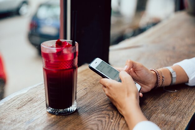 female hands typing a message on mobile phone, sitting for the table with fruit cocktail