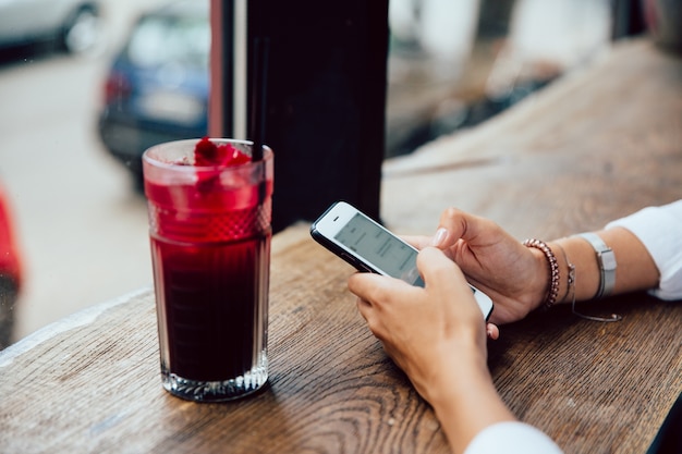 female hands typing a message on mobile phone, sitting for the table with fruit cocktail