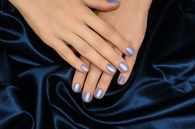 Female hands with blue nail design. Blue glitter nail polish manicure. Woman hands on blue fabric background