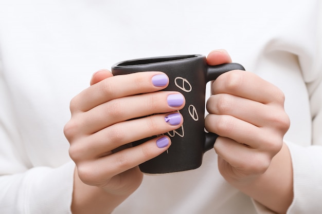 Женские руки с фиолетовым дизайном ногтя держа черную чашку.