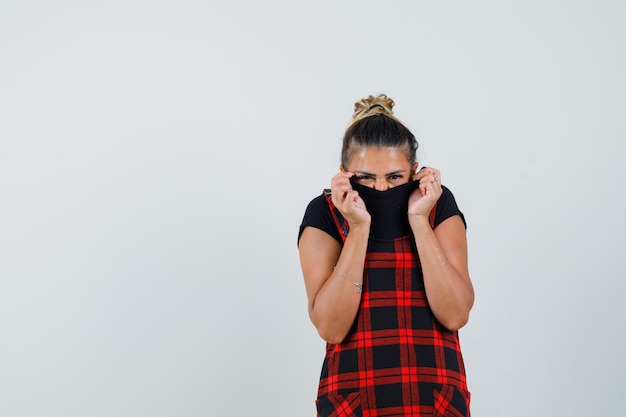 Free photo female hiding face behind her collar in pinafore dress and looking scared , front view.