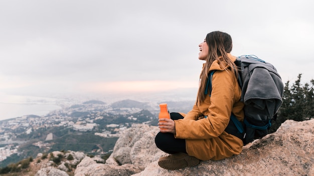 A female hiker sitting on the top of mountain holding water bottle in hand overlooking the view