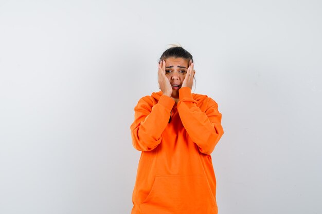 Female holding hands on cheeks in orange hoodie and looking wistful 