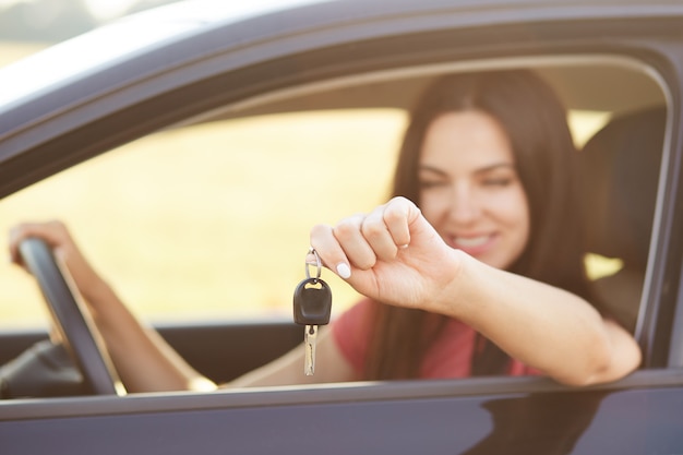 Female holds key while sits in luxury automobile, glad to recieve expensive present from relatives, focus on keys