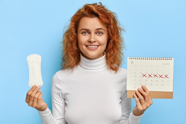 Free photo female hygiene concept. redhead smiling woman holds clean period sanitary napkin pad and menstruation calendar