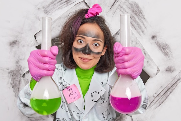 Женщина-исследователь лаборатории держит две колбы с разноцветной жидкостью, имеет грязное лицо после взрыва, носит медицинский халат и перчатки показывает результаты своего эксперимента в лаборатории.