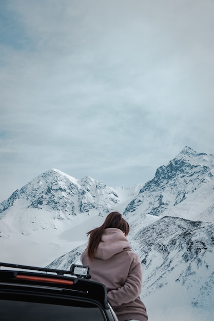 무료 사진 놀라운 눈 덮인 바위 산과 흐린 하늘 앞에서 검은 차량에 기대어 여성