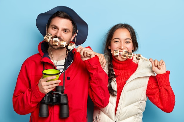 Female and male tourists eat tasty marshmallow roasted on bonfire, spend pastime in nature, like traveling and having adventures, wear casual clothes