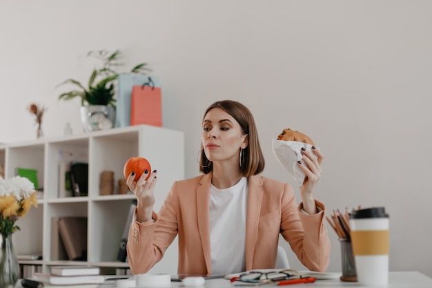 Female manager posing in white office. Lady looks at apple reluctantly, wanting to eat burger.