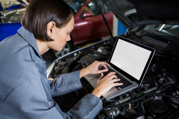 Female mechanic examining car engine with help of laptop