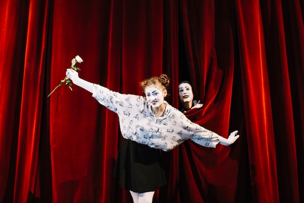 Female mime holding white rose performing on stage with male mime peeking behind the curtain