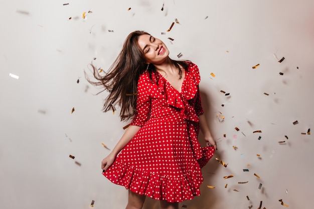 Free photo female model closed her eyes from pleasure and dancing with smile on white wall with falling confetti. photo of lady in red dress.