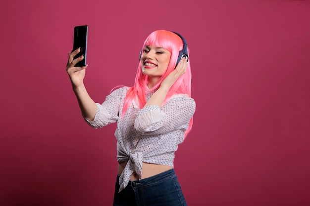 Бесплатное фото Женская модель с розовыми волосами фотографирует или записывает видео на смартфон, слушает музыку в наушниках. используйте мобильный телефон, чтобы делать фотографии и слушать песни на гарнитуре для развлечения.