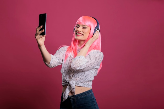 スマートフォンで写真を撮ったりビデオを録画したり、ヘッドフォンで音楽を聴いたりする、ピンクの髪の女性モデル。携帯電話を使用して写真を撮り、エンターテイメント用のヘッドセットで曲を楽しむ。