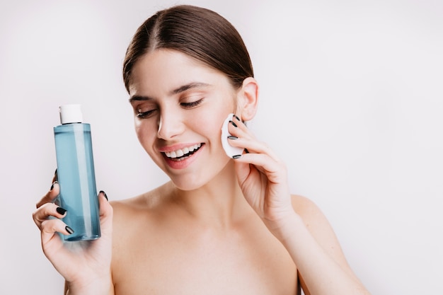 Female model with snow-white smile and without make-up poses on white wall, demonstrating beneficial properties of micellar water.