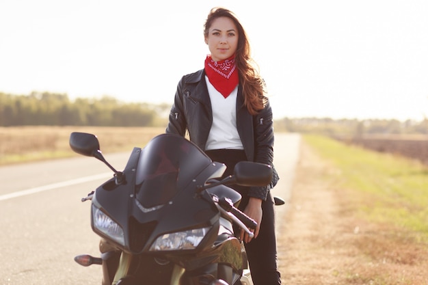 Female motocross racer dressed in black leather jacket, poses on her motorcycle, has adventure in countryside, likes risky sport