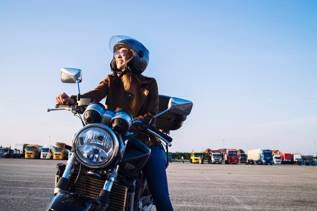 Женщина-мотоциклист в кожаной куртке и шлеме сидит на ретро-мотоцикле и улыбается