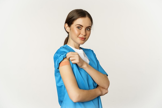 女性看護師、医師の医療従事者はワクチン接種を受け、covid-19ワクチン接種後にパッチで肩を見せ、満足そうに笑い、白い背景の上に立った。