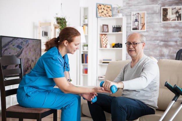 Female nurse doing physiotherapy with senior man in nursing home using dumbbells.