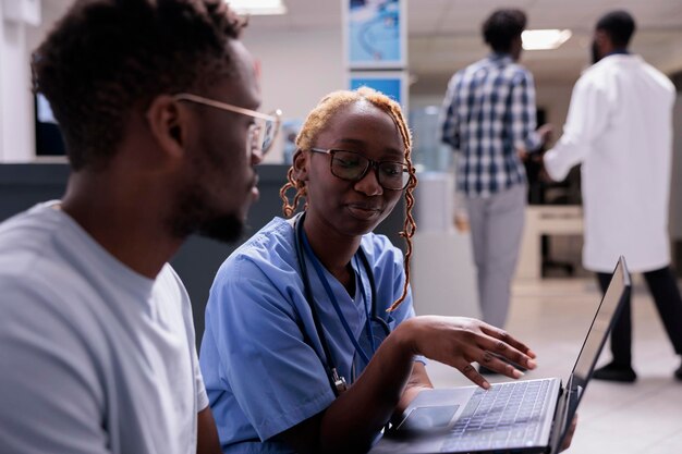 Female nurse using laptop with patient at consultation in waiting area, showing diagnosis results on computer and giving healthcare treatment. Talking to man about disease and medical recovery.