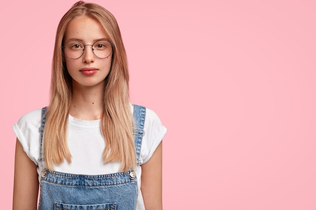 Female office worker relaxes after long day at work, dressed casually, expresses confidence, wears round glasses and dungarees, stands alone against pink wall with copyspace