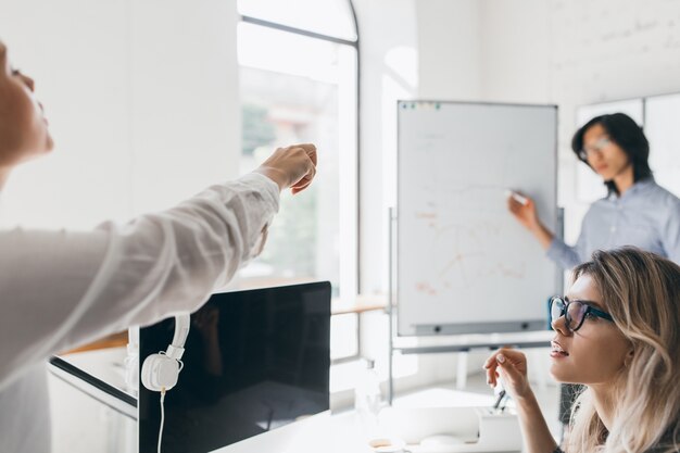 Female office worker in white blouse pointing finger at board during meeting with colleagues