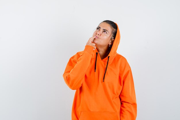 Female in orange hoodie keeping finger on lips and looking pensive 